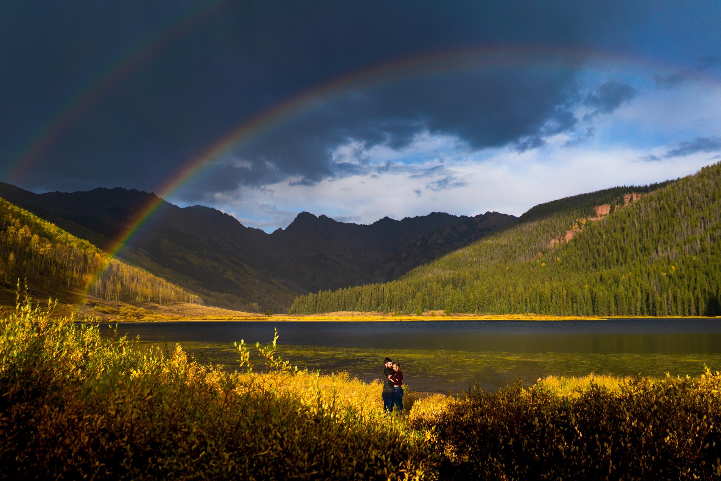 Engaged couple embraces for a portrait during a rainstorm with a double rainbow behind them arching over Piney Lake, Engagement Session, Engagement Photos, Engagement Photos Inspiration, Engagement Photography, Engagement Photographer, Fall Engagement Photos, Mountain Engagement Photos, Piney River Ranch engagement photos, Vail engagement session, Vail engagement photos, Vail engagement photography, Vail engagement photographer, Vail engagement inspiration, Colorado engagement session, Colorado engagement photos, Colorado engagement photography, Colorado engagement photographer, Colorado engagement inspiration